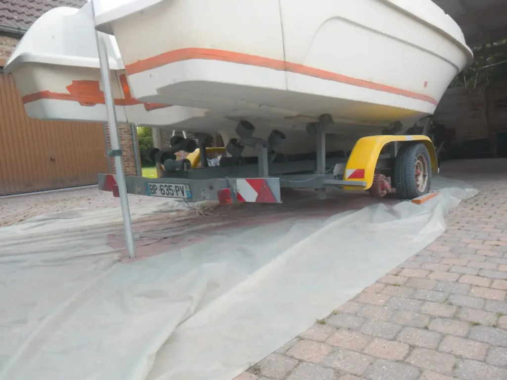 Vue arrière d'un bateau à moteur blanc de marque 'Sea-Serpent' reposant sur une remorque routière dans une allée pavée. La remorque est galvanisée avec des garde-boue jaunes et une bâche en plastique protège le sol des éclaboussures de peinture rouge.