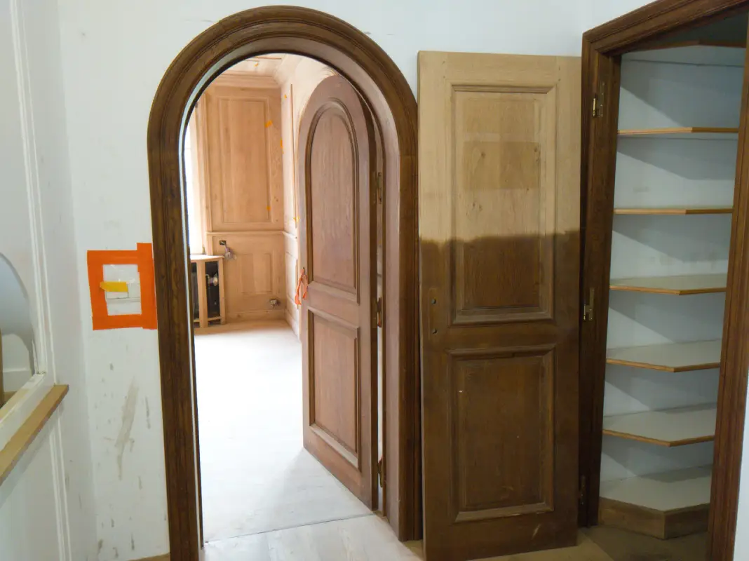 Une photo d'un couloir en rénovation. Une grande arche en chêne encadre une porte ouverte menant à une pièce entièrement lambrissée de bois clair. À droite, une porte de style identique est posée, montrant une délimitation nette entre la partie supérieure décapée (bois clair) et la partie inférieure d'origine (bois foncé). Plus à droite, un placard encastré avec des étagères blanches et des côtés en chêne est ouvert. Les murs blancs sont marqués de taches et d'un carré d'adhésif orange. Le sol est en plancher brut.